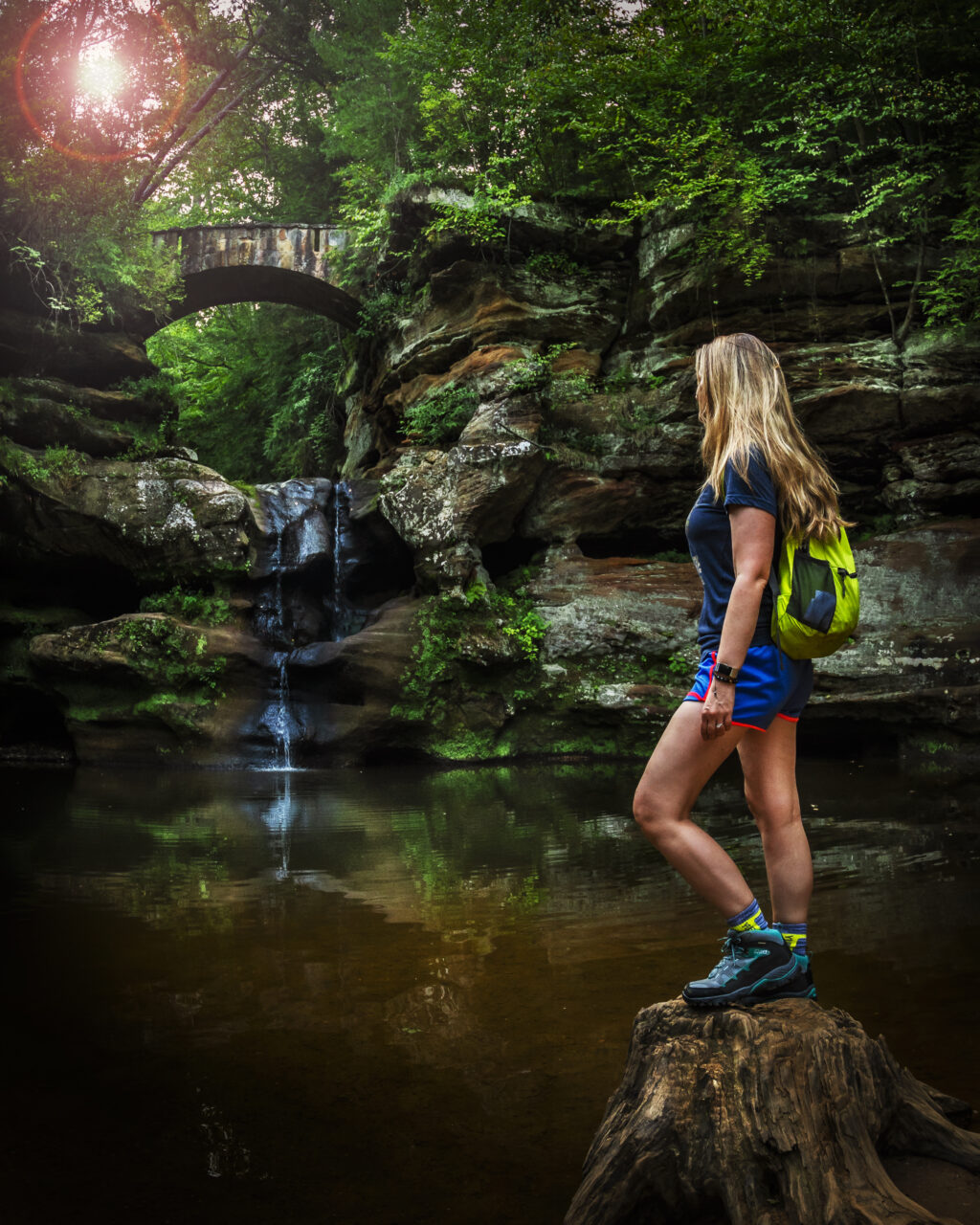 Woman standing on a tree stump with a waterfall in the background in a wooded area.