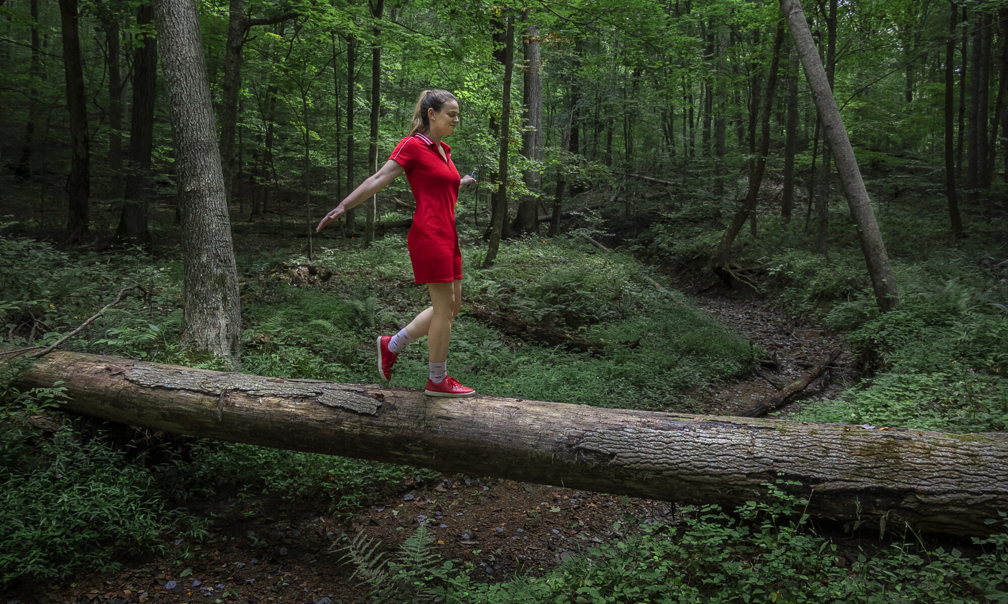 Woman in red dress walking across a fallen tree over a creek bed in the forest.