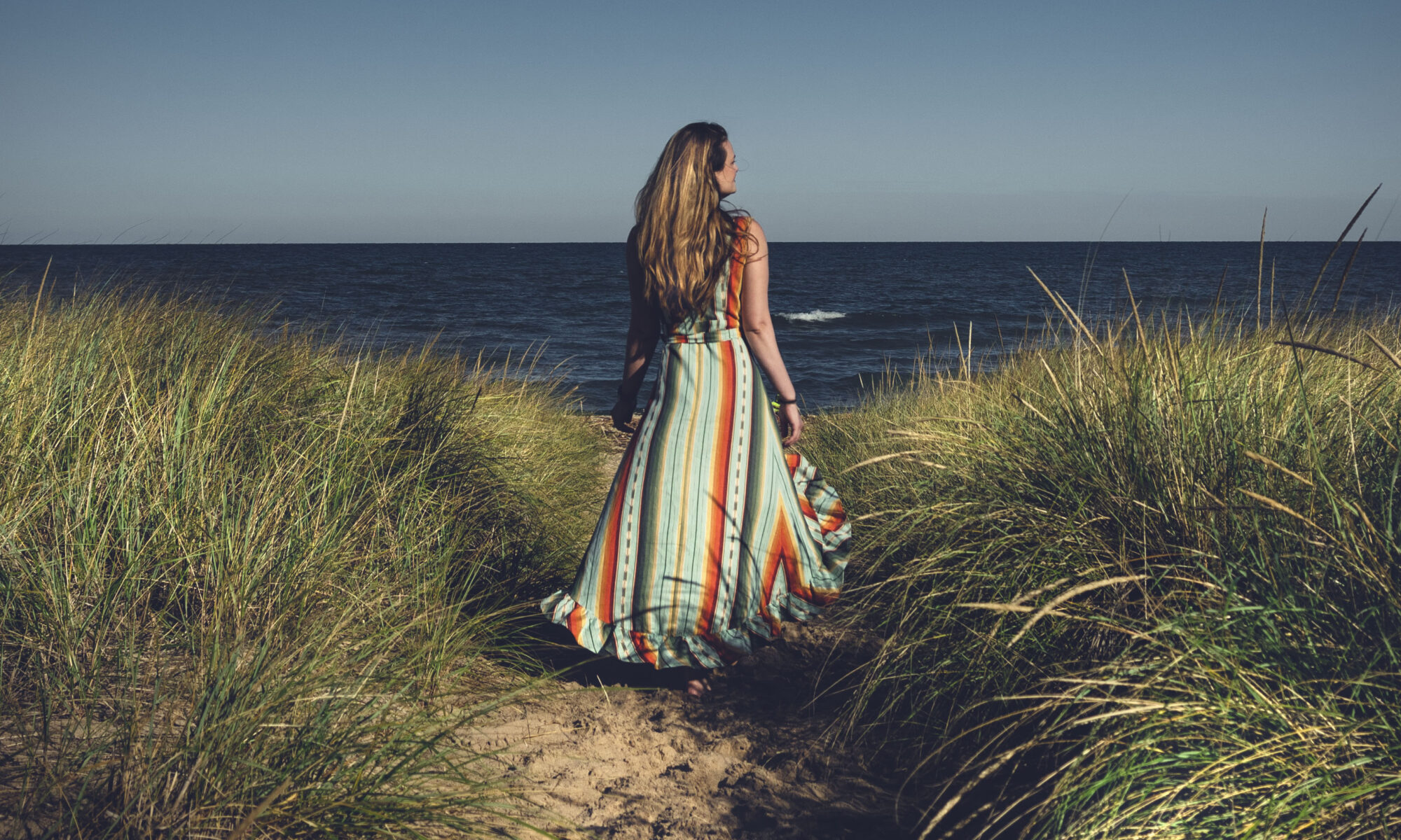 A woman in a colorful dress walks a sandy path in a grassy plain with Lake Michigan in the background.