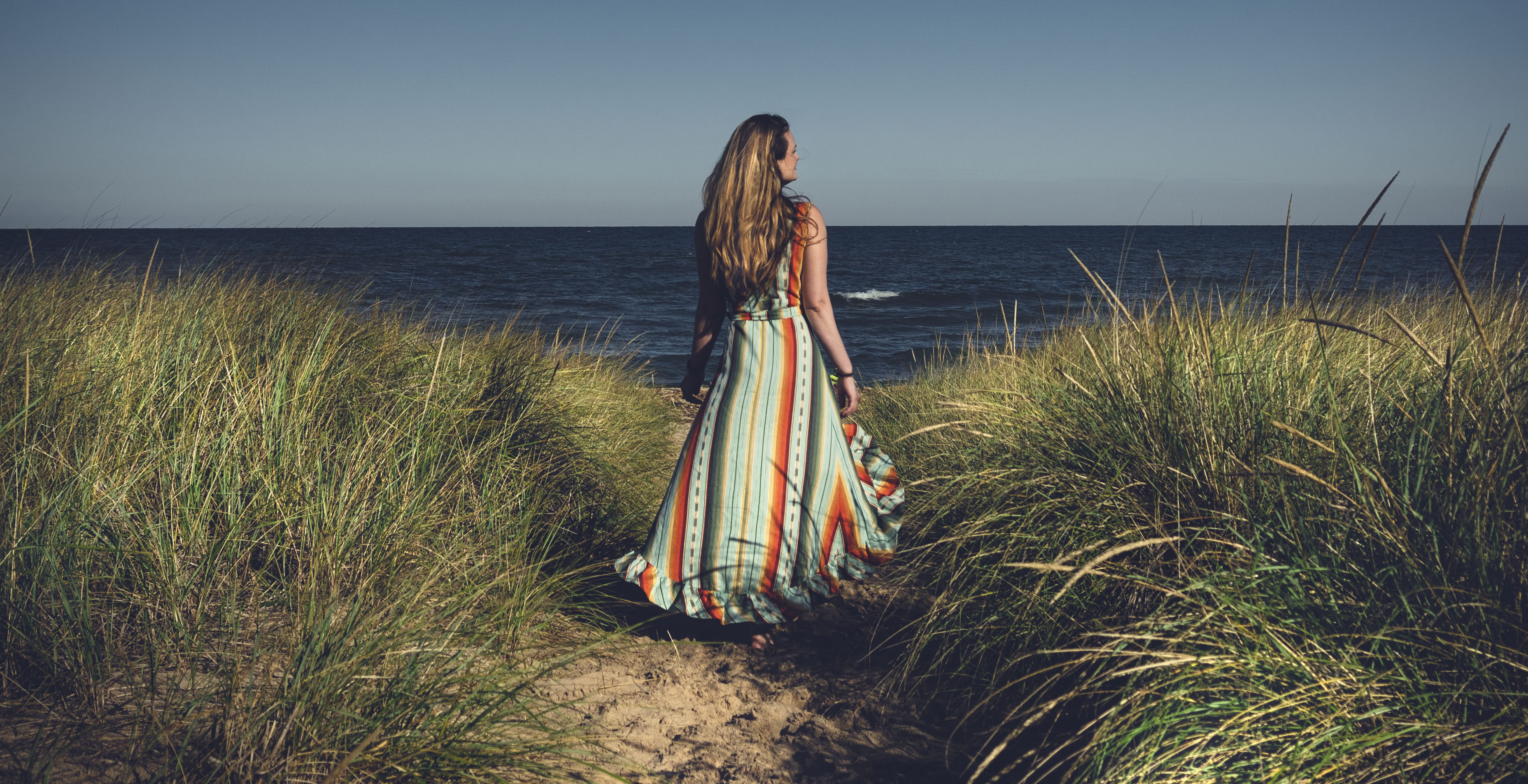 A woman in a colorful dress walks a sandy path in a grassy plain with Lake Michigan in the background.