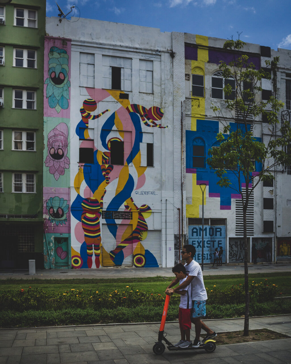 Children ride a scooter past colorful murals on a row of three story building on Boulevard Olimpico, Brazil.