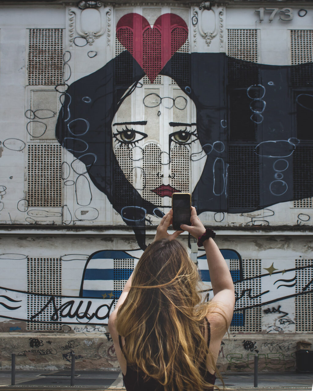 A woman holds up her phone to photograph a mural of a woman's face on a three story building in Rio de Janeiro.