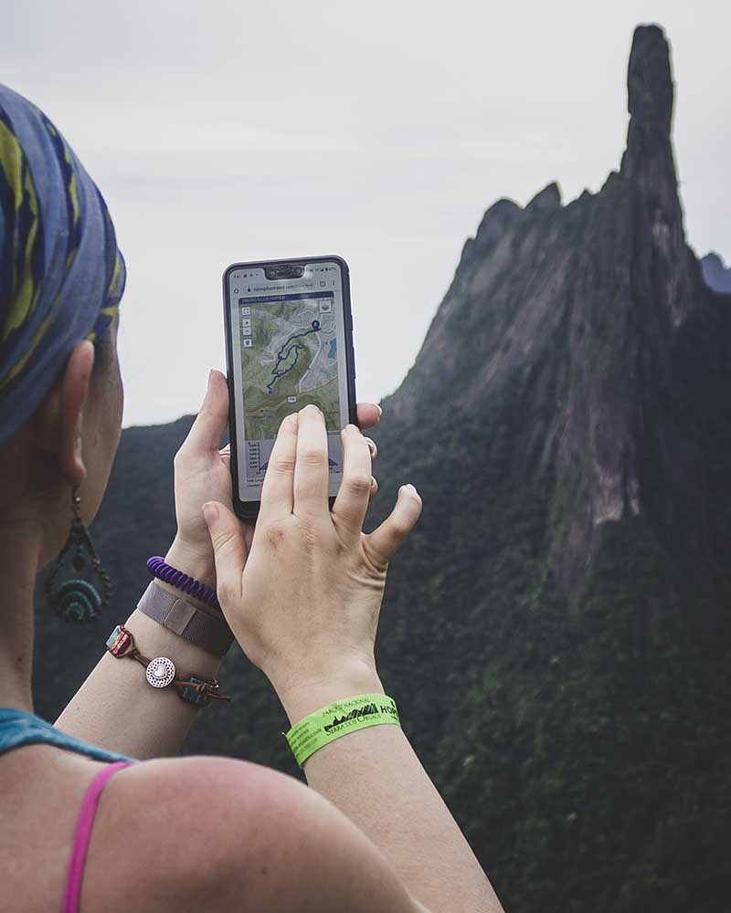 Woman holding smartphone displaying interactive hiking illustrated map with Dedo de Deus mountain in background.