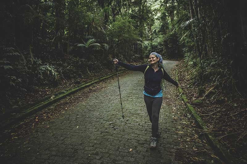 Woman hiker with arms outstretched walking on brick road surrounded by lush forest.