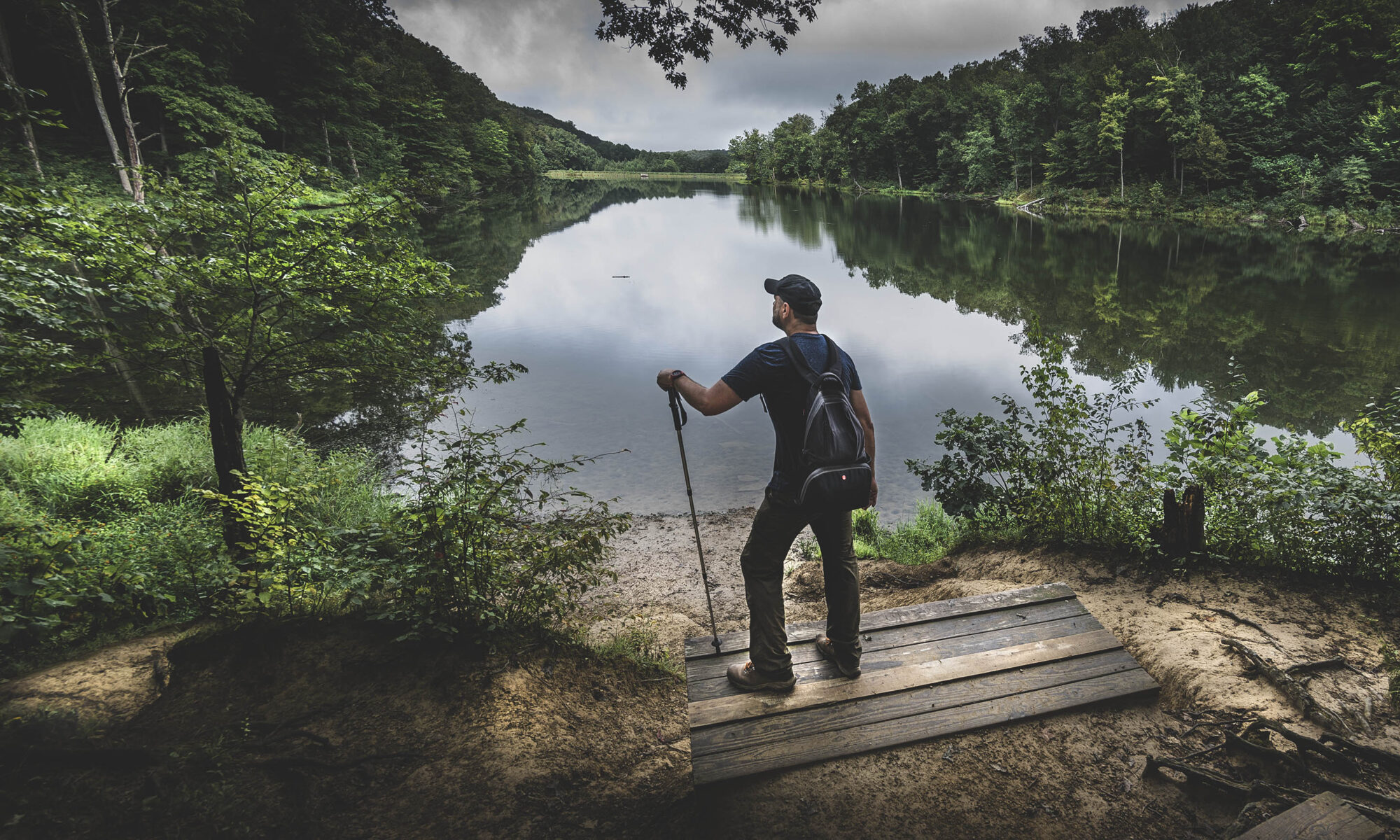 Male hiker looking out over a lake surrounded by trees and greenery.