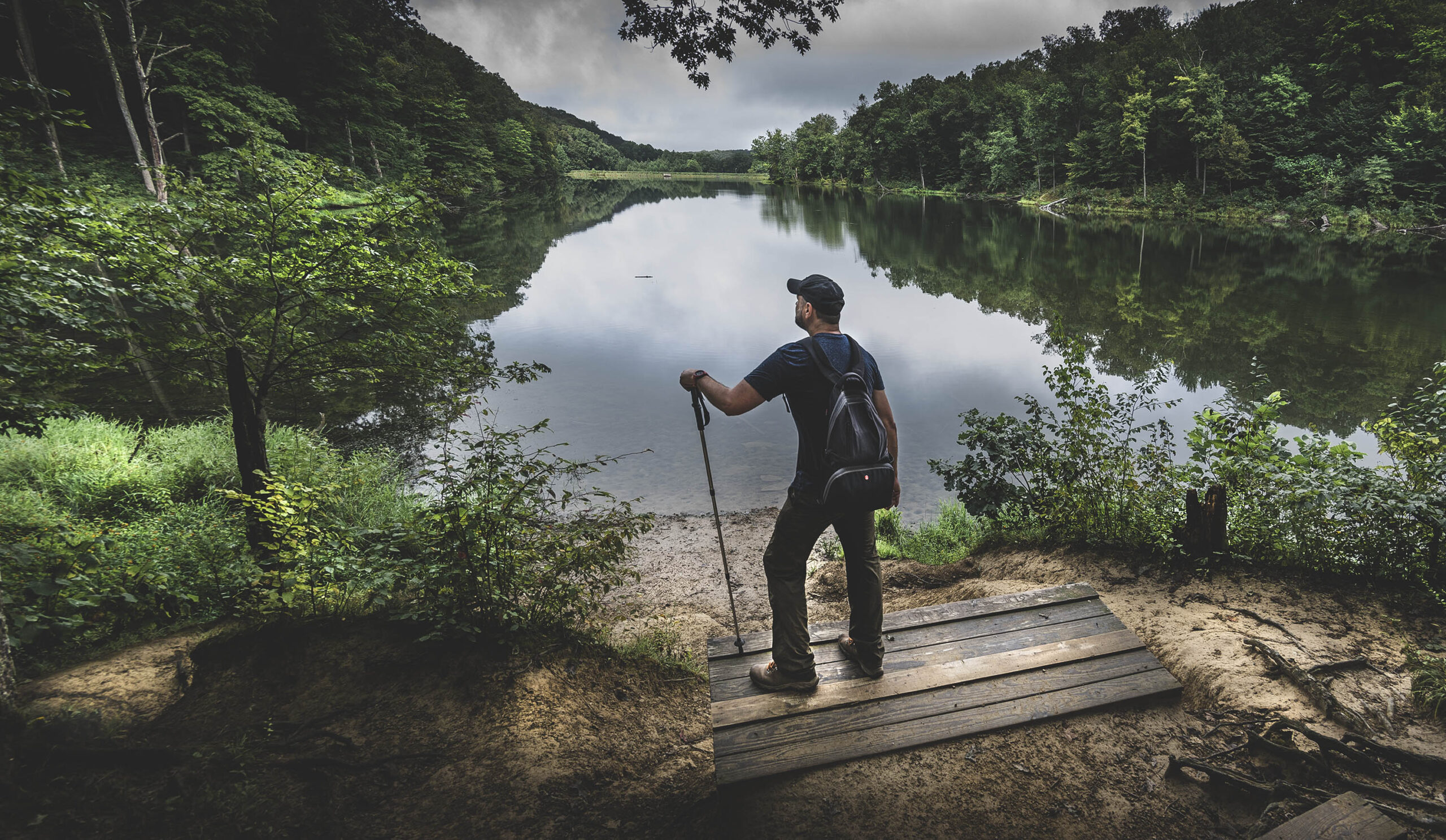 Male hiker looking out over a lake surrounded by trees and greenery.